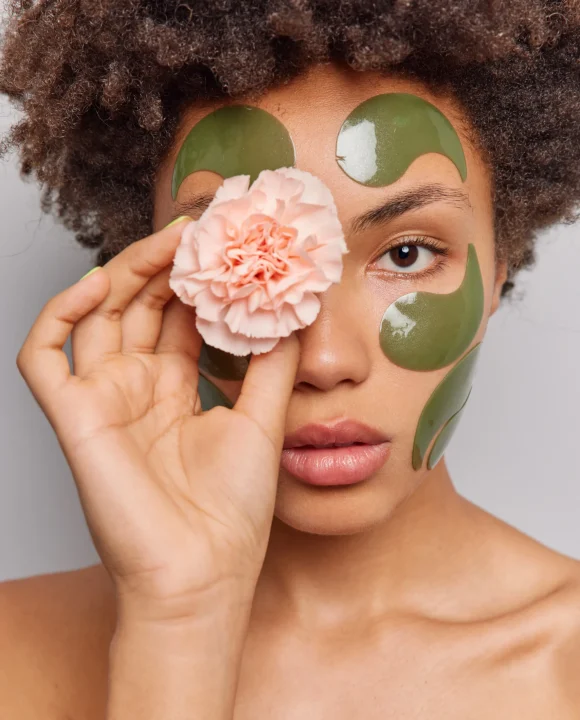 woman-uses-natural-beauty-products-holds-flower-eye-applies-collagen-green-patches-face-stands-shirtless-poses-indoor